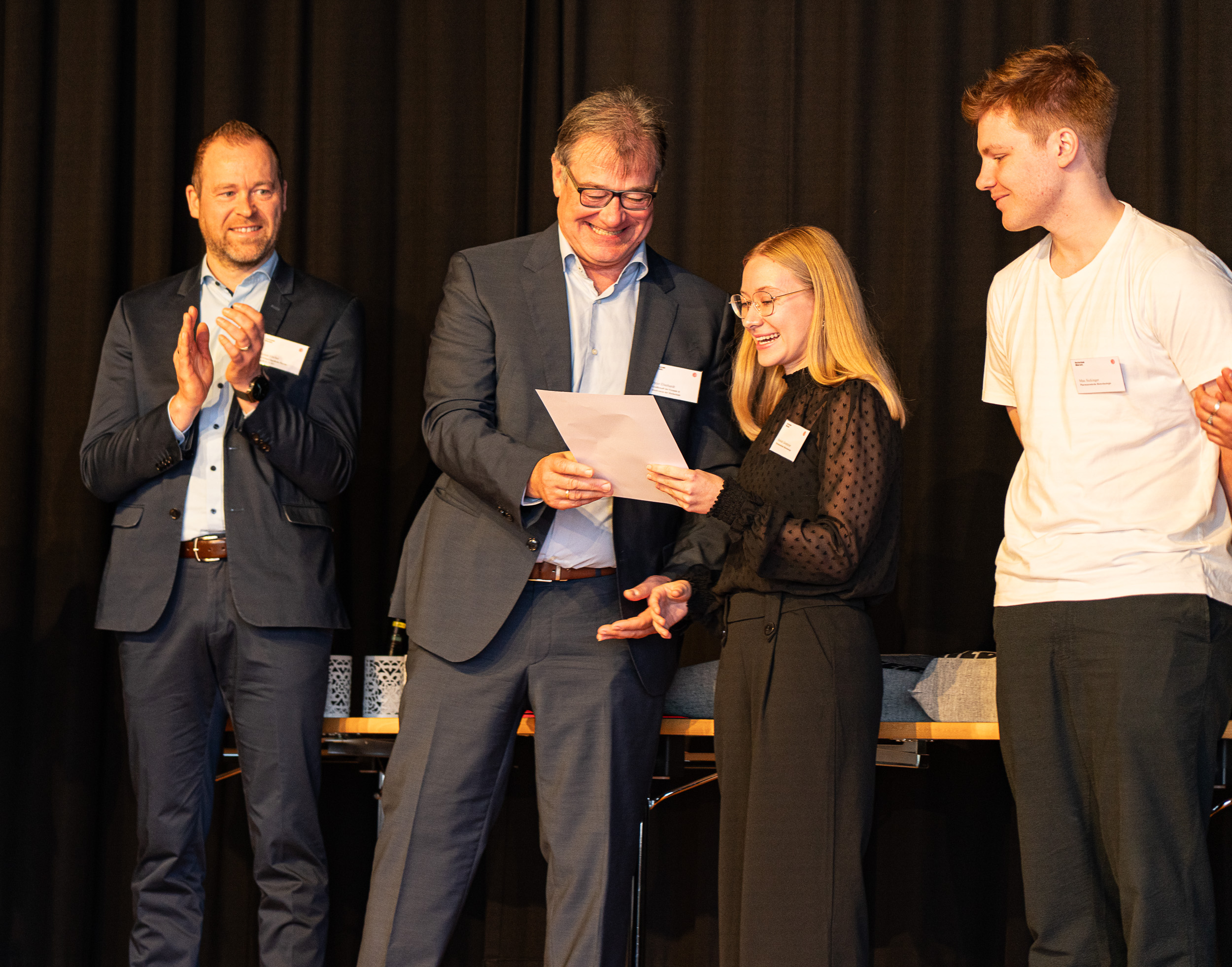 Zwei Männer und eine junge Frau und ein junger Mann stehen auf einer Bühne und bekommen Preise übergeben