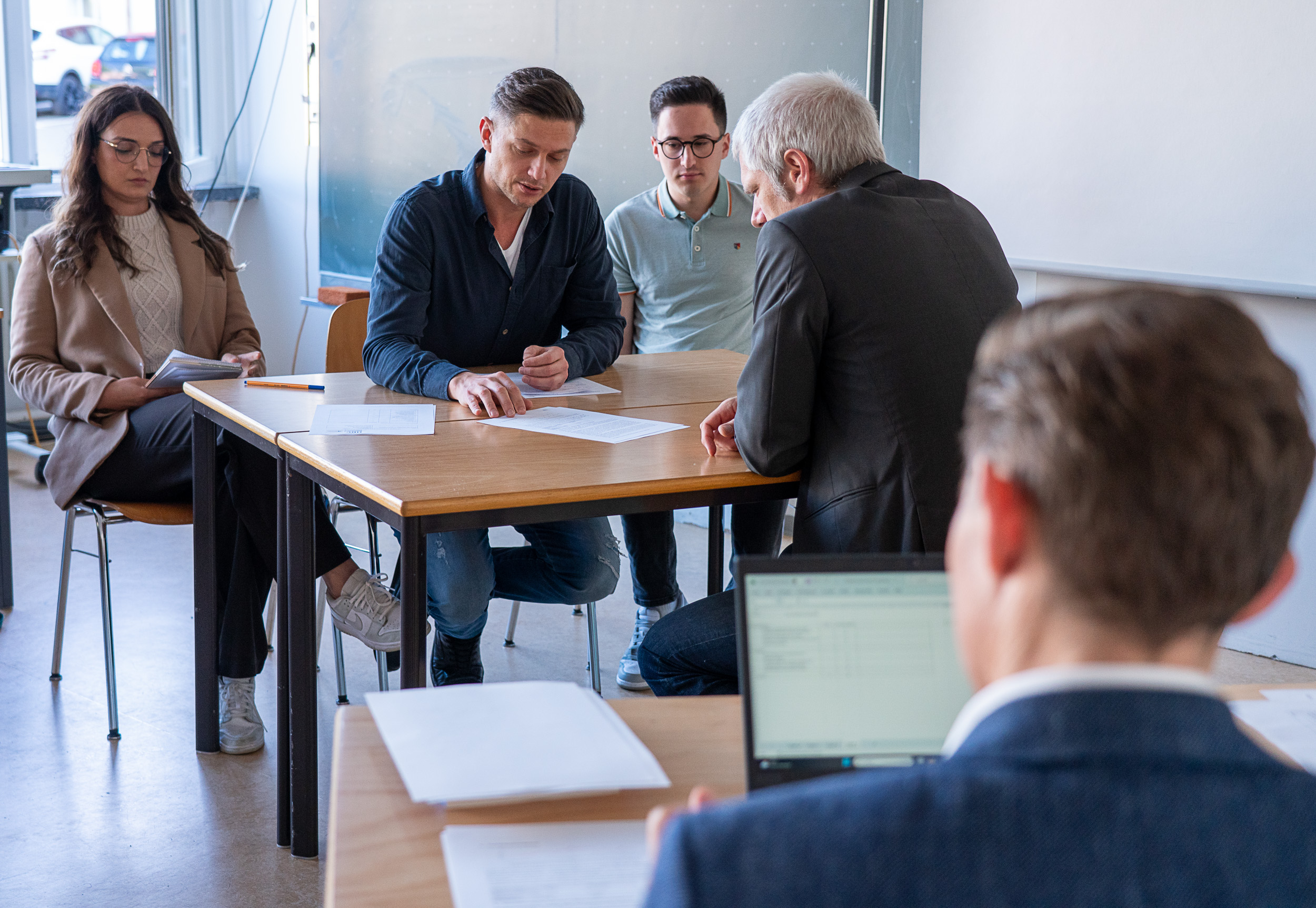 Vier Personen sitzen an einem Tisch und werden von einem Mann geprüft