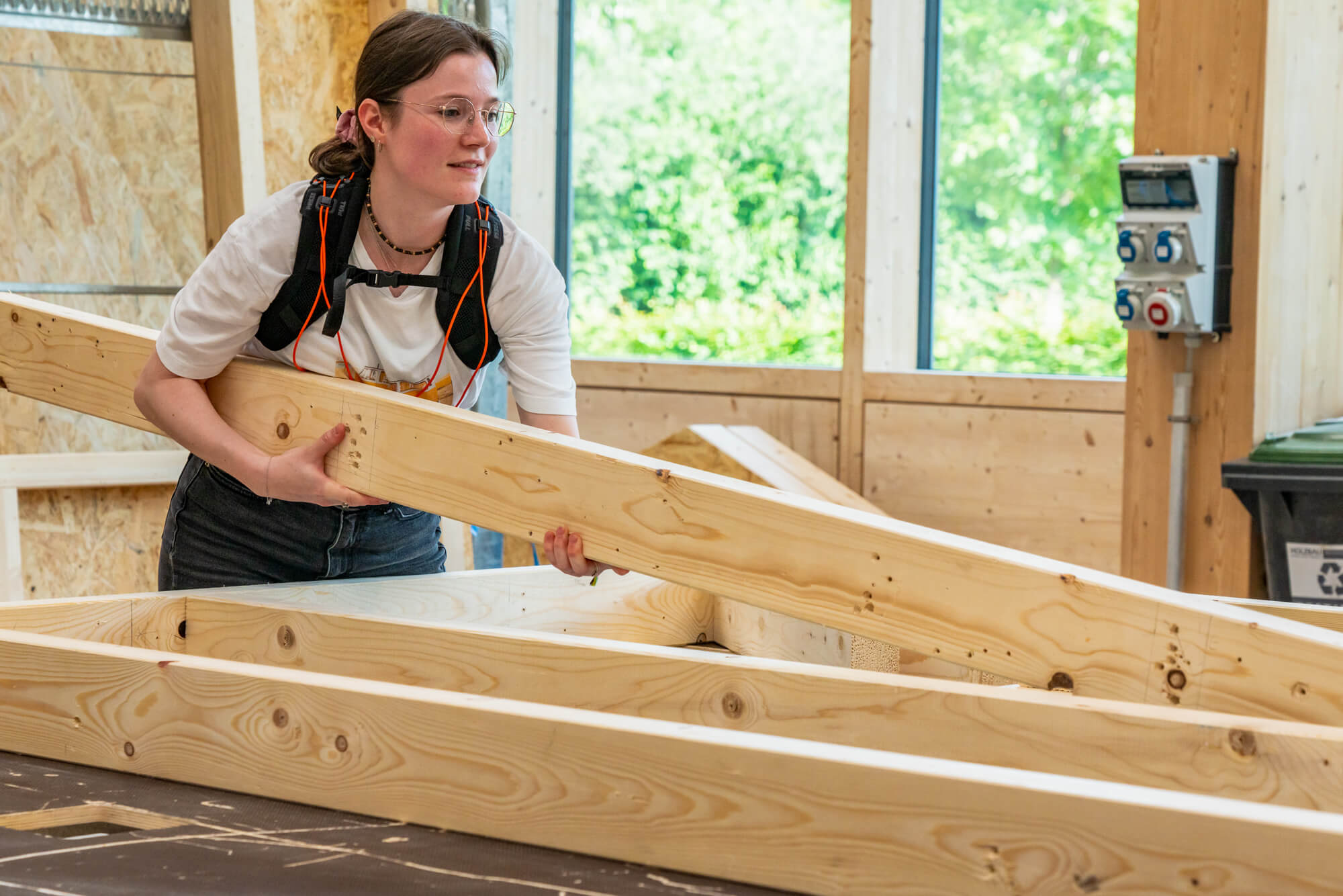 Eine Studierende mit Exoskelett trägt einen schweren Holzbalken