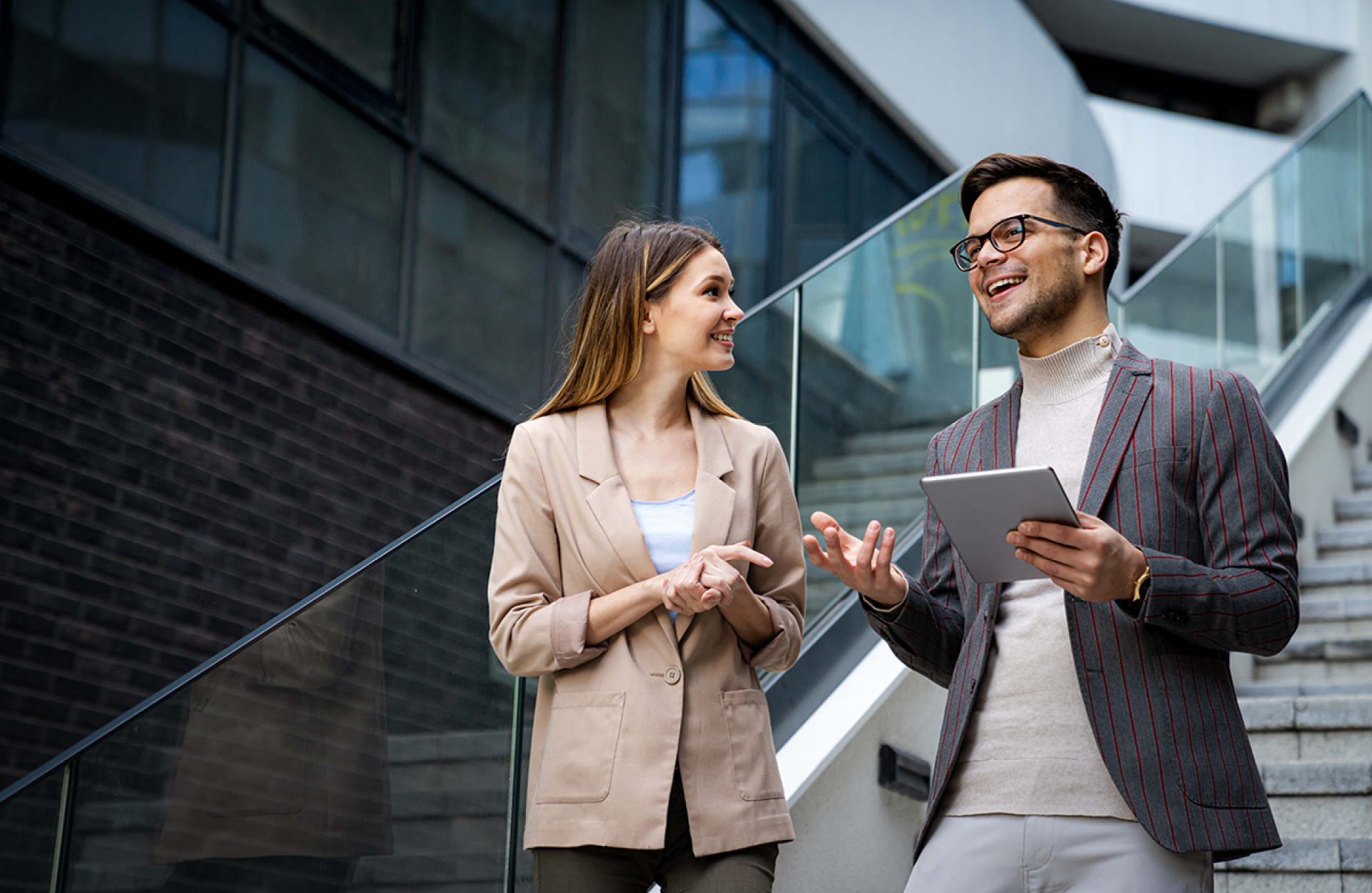 Eine Frau und ein Mann mit Tablet, beide im Business Look, stehen auf einer Treppe in einem modernen Unternehmen und unterhalten sich
