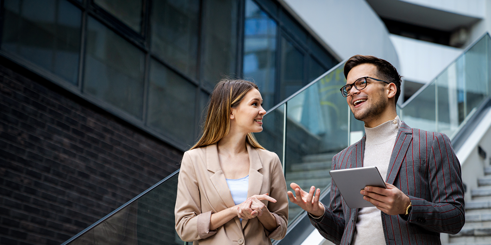 Eine Frau und ein Mann mit Tablet, beide im Business Look, stehen auf einer Treppe in einem modernen Unternehmen und unterhalten sich