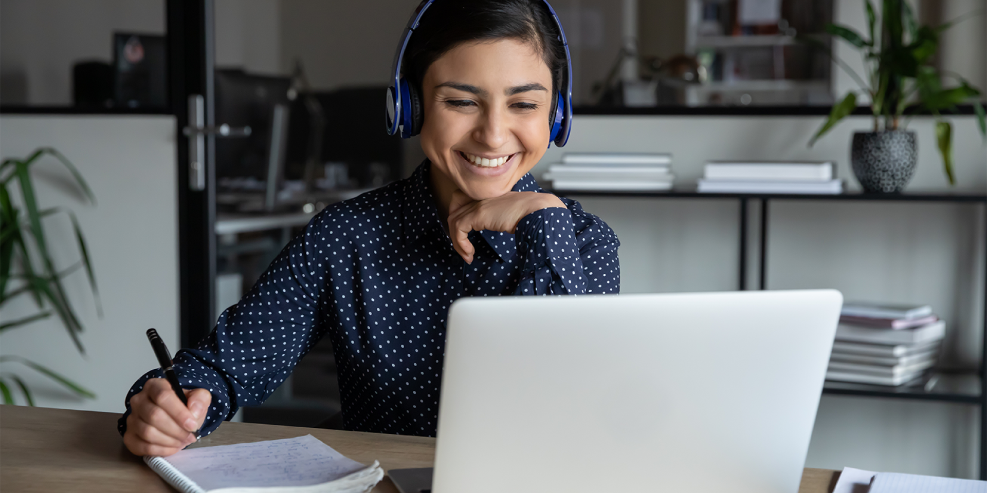 Junge Frau sitzt lächelnd it Headset und Schreibset vor einem Laptop in einem modernen Büro