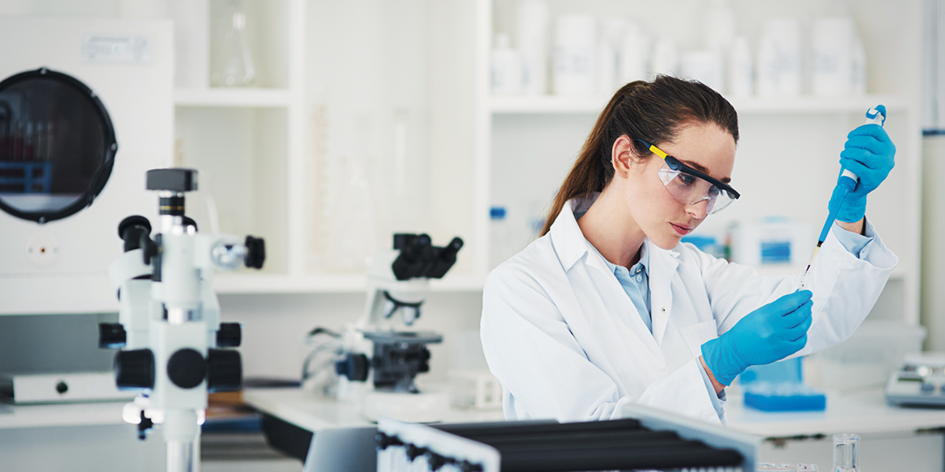 Eine Frau im weißen Kittel mit Schutzbrille und blauen Handschuhen steht in einem modernen Labor und arbeitet mit einer Pipette