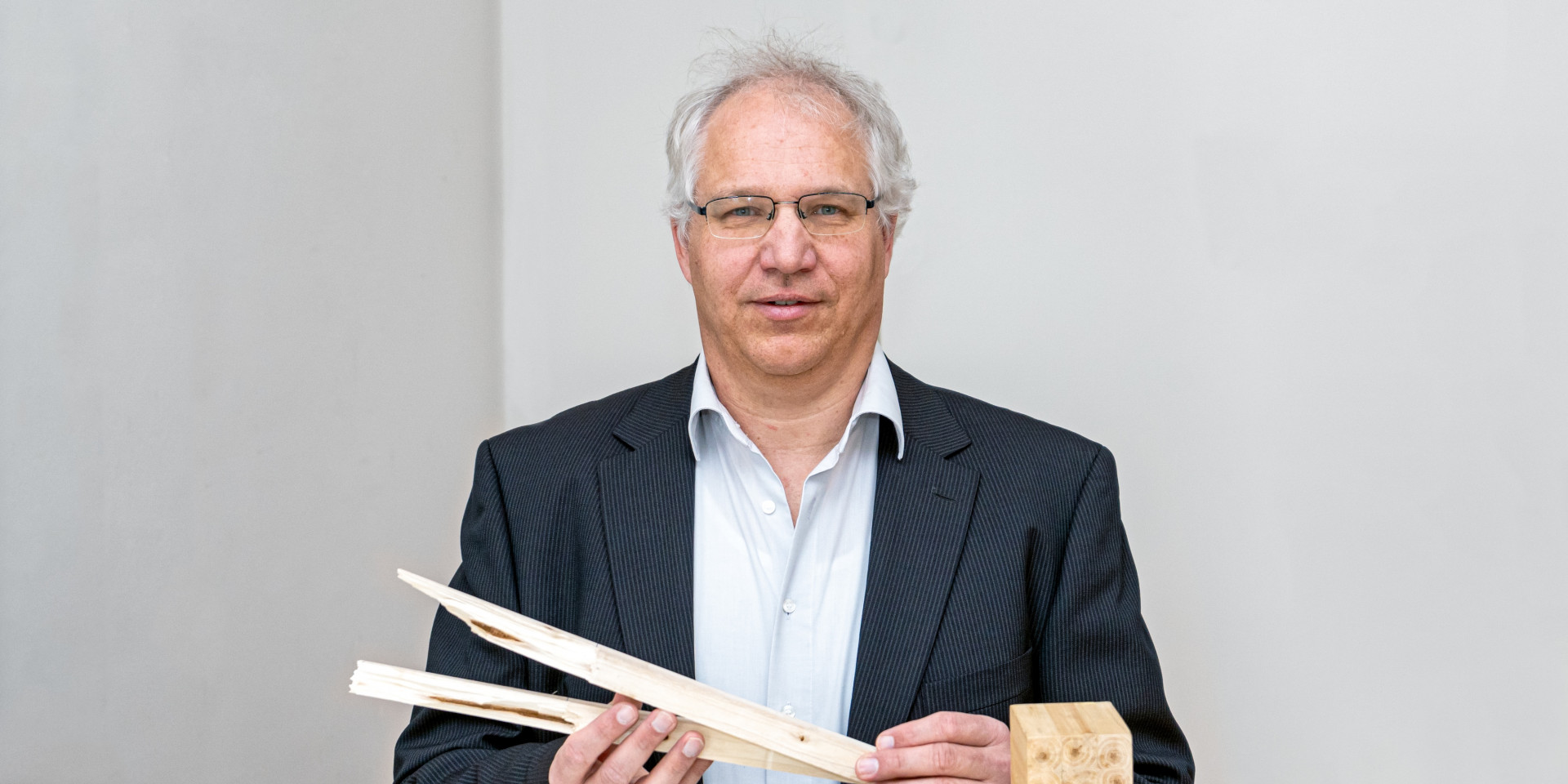 Mann im Anzug steht vor einer weißen Wand und hält Holz in der Hand