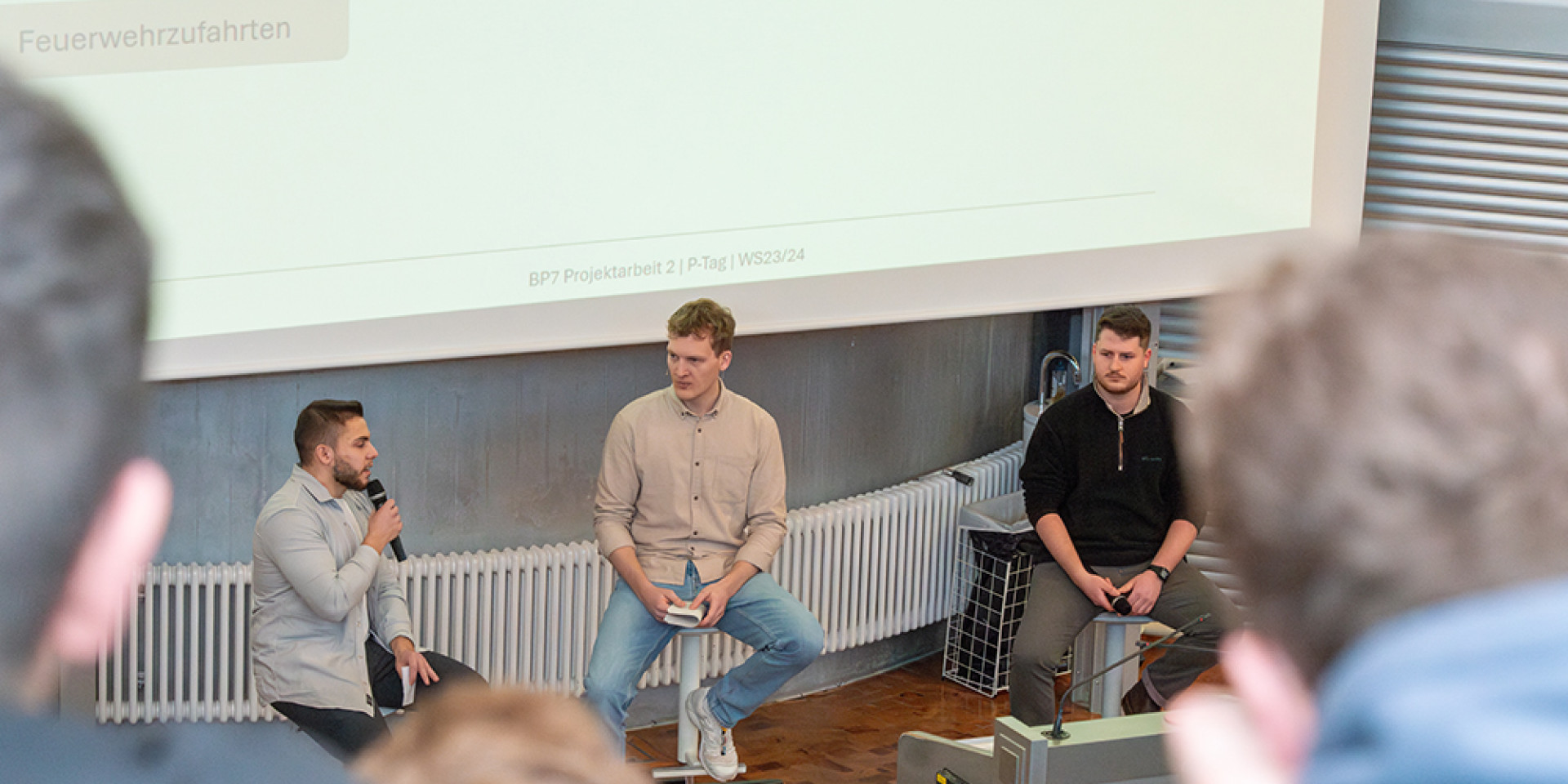 Drei Studenten sitzen im Vorlesungsraum vor der Leinwand und präsentieren