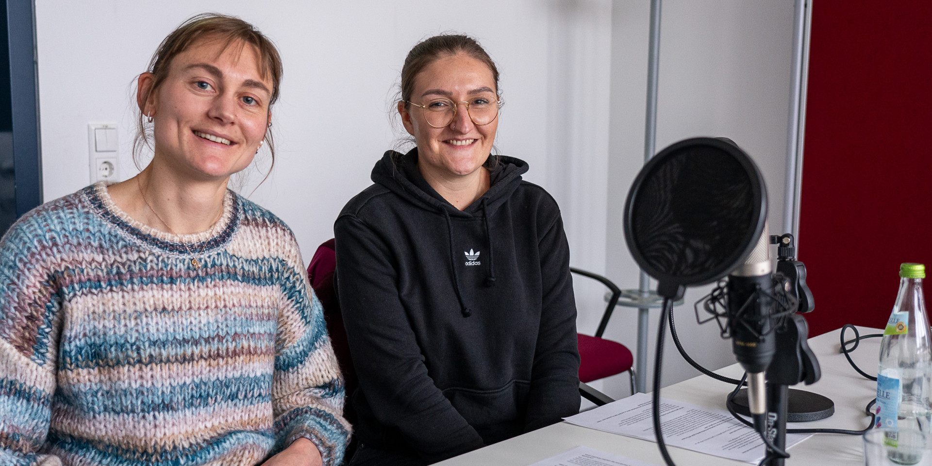 Zwei Frauen sitzen an einem Tisch bei einer Podcastaufnahme und lächeln in die Kamera