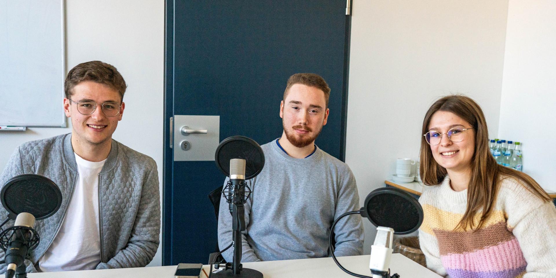 Drei Studierende sitzen mit Mikrofonen um einen Tisch