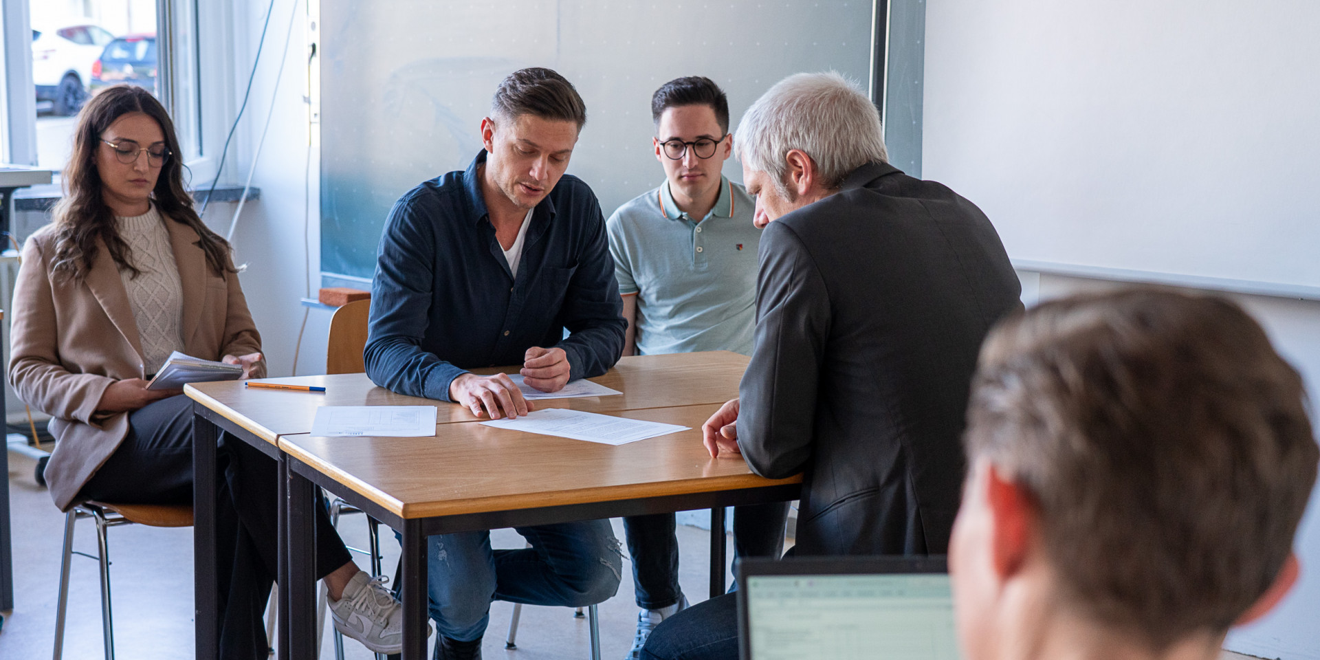 Vier Personen sitzen an einem Tisch und werden von einem Mann geprüft