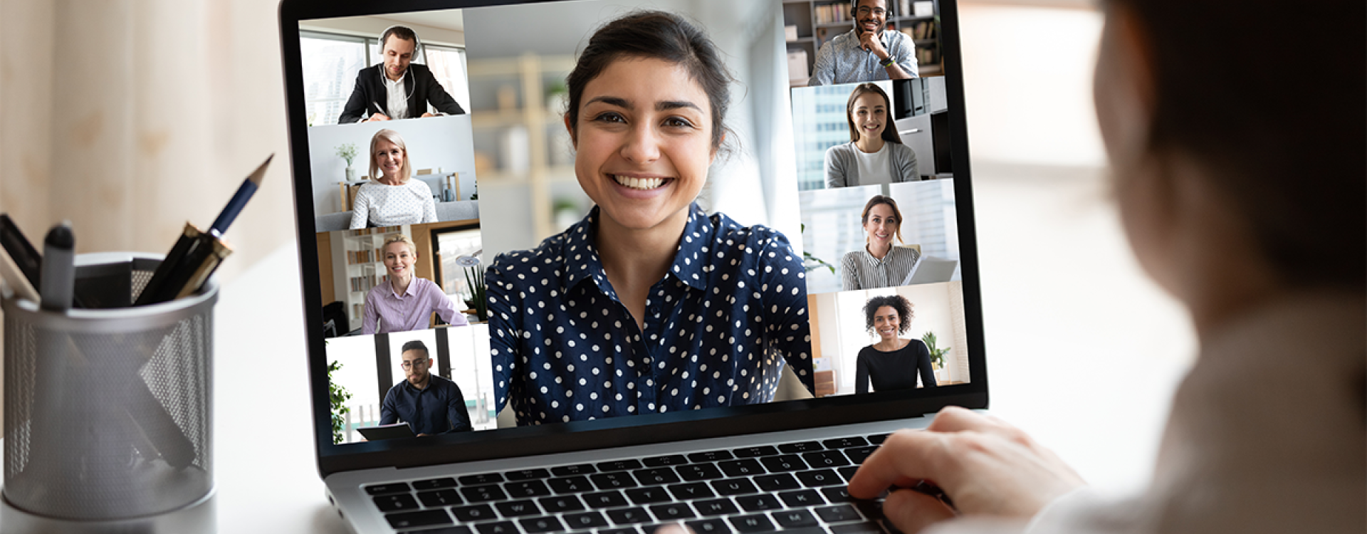 Frau mit weißer Bluse sitzt vor einem Laptop am Schreibtisch und nimmt an einem Online-Meeting teil, dessen Teilnehmer auf dem Bildschirm zu sehen sind