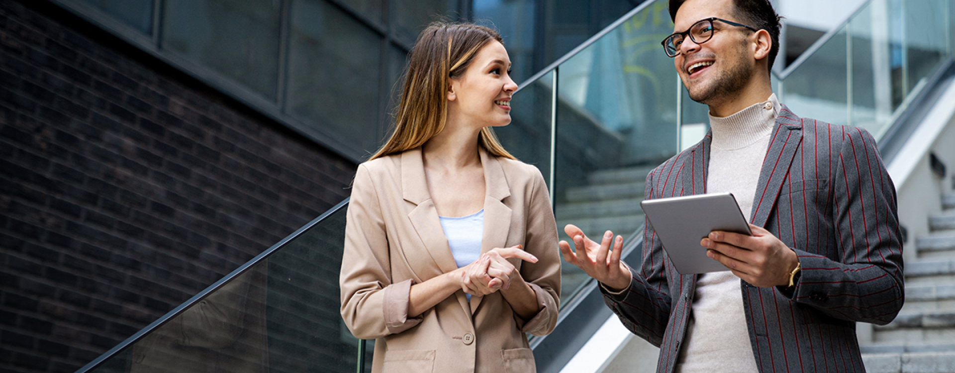 Eine Frau und ein Mann mit Tablet in der Hand, beide im Business Look, stehen auf einer Treppe in einem modernen Unternehmen und unterhalten sich,