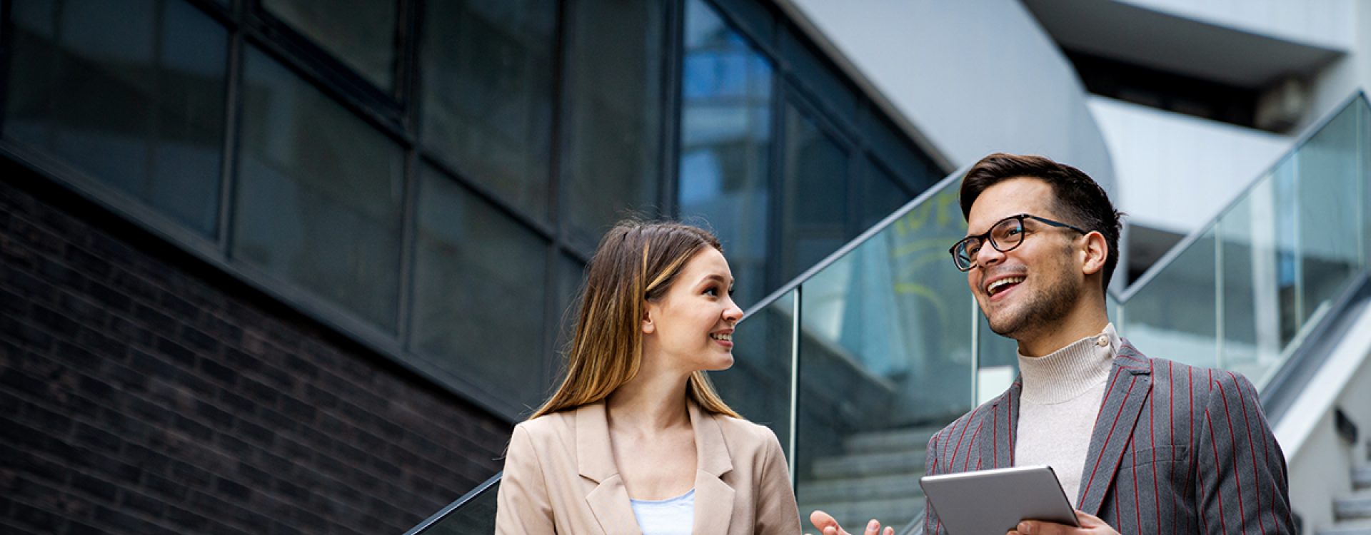 Eine Frau und ein Mann mit Tablet, beide im Business Look, stehen auf einer Treppe in einem modernen Unternehmen und unterhalten sich