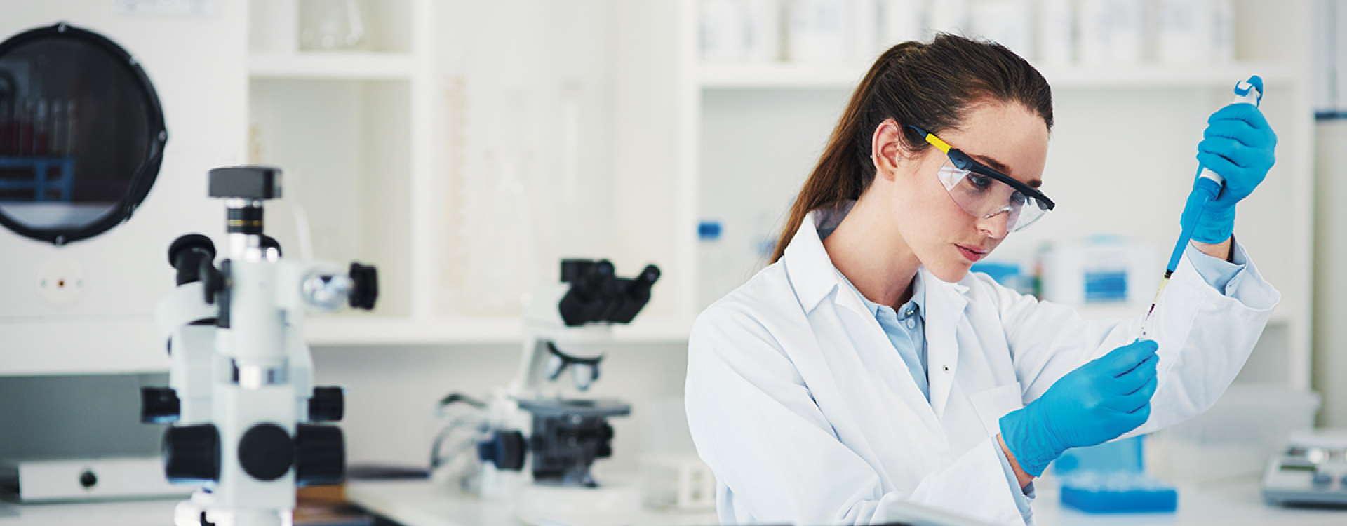Eine Frau im weißen Kittel mit Schutzbrille und blauen Handschuhen steht in einem modernen Labor und arbeitet mit einer Pipette