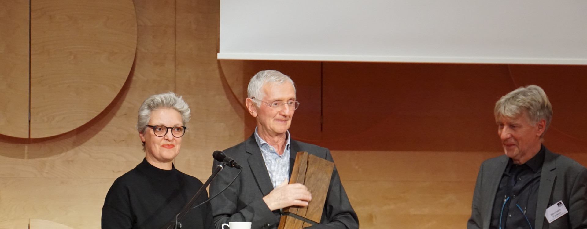 Drei Personen vor Stehpult auf einer Bühne