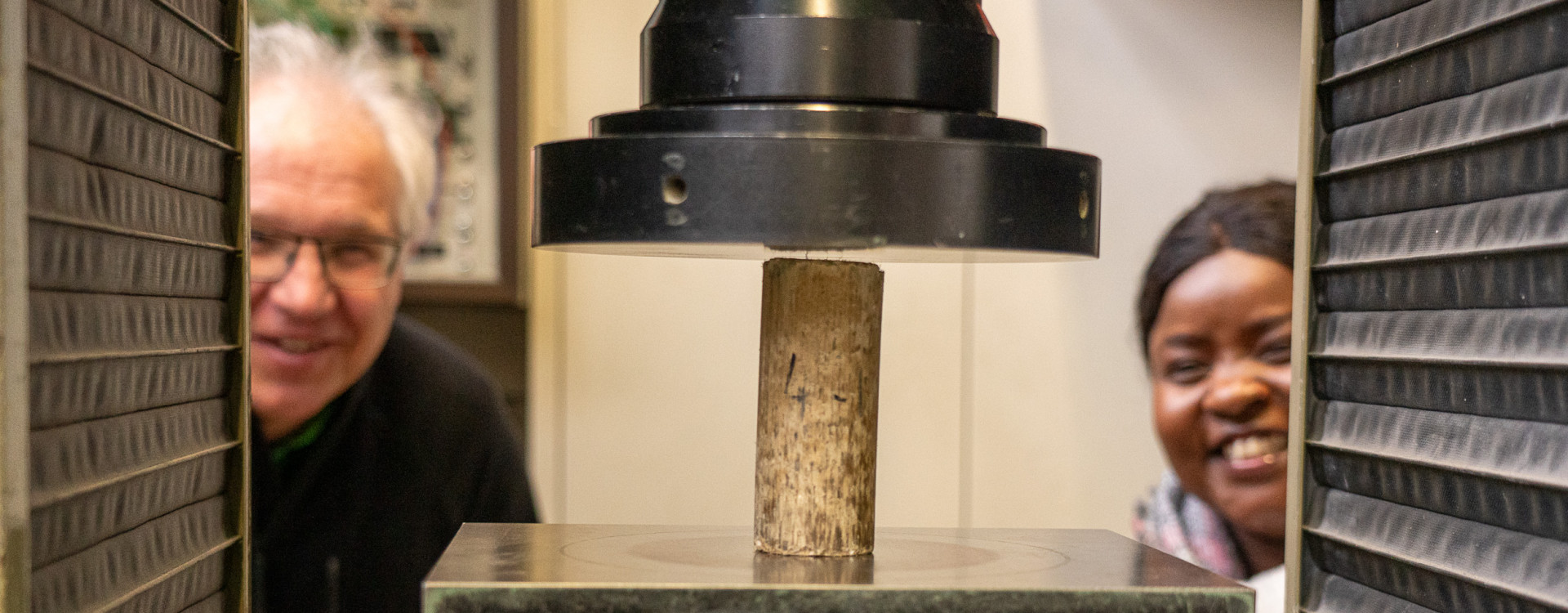 Zwei Personen schauen sich einen Lehmstein ein, der von einer Maschine zusammengepresst wird