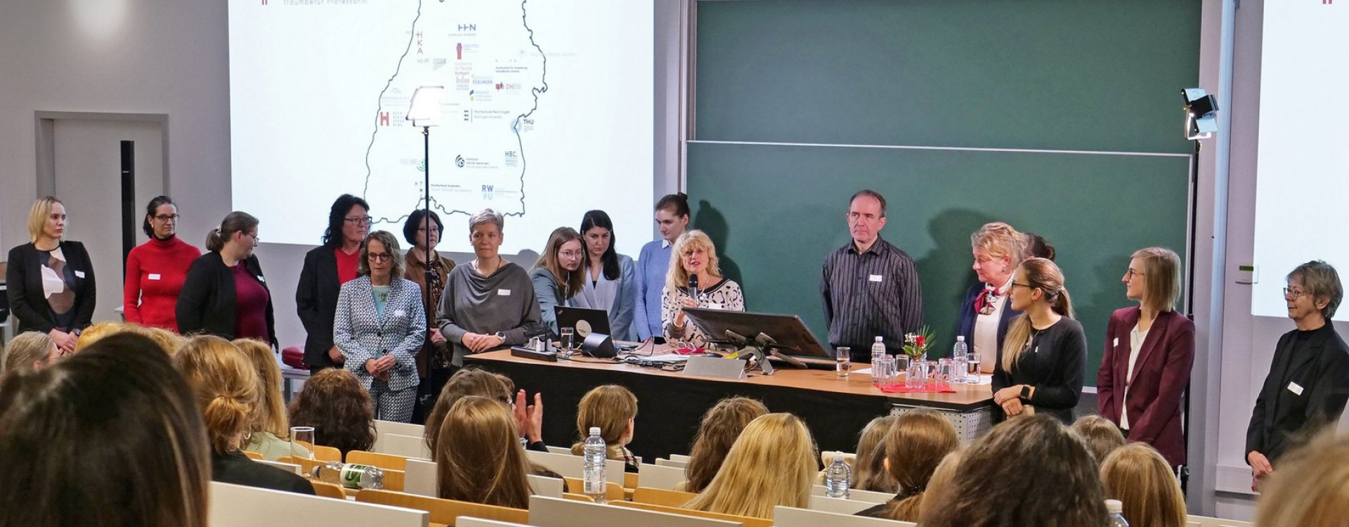 Viele Menschen stehen in Hörsaal vor Tafel