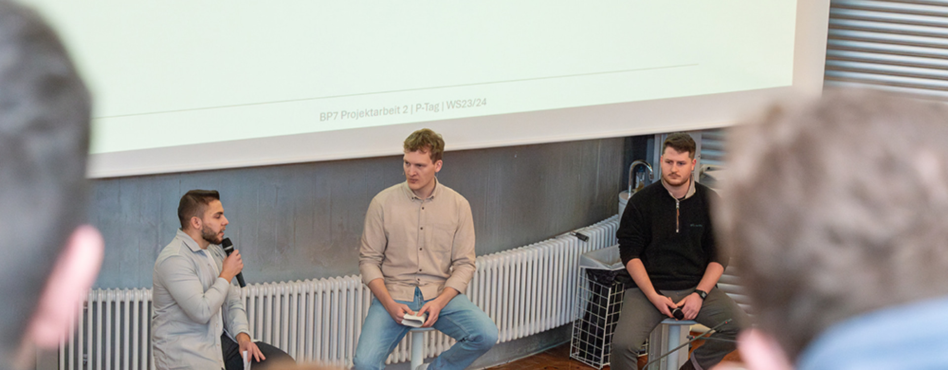 Drei Studenten sitzen im Vorlesungsraum vor der Leinwand und präsentieren