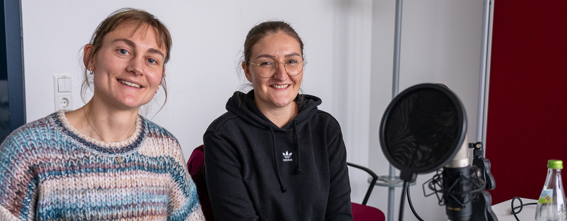 Zwei Frauen sitzen an einem Tisch bei einer Podcastaufnahme und lächeln in die Kamera