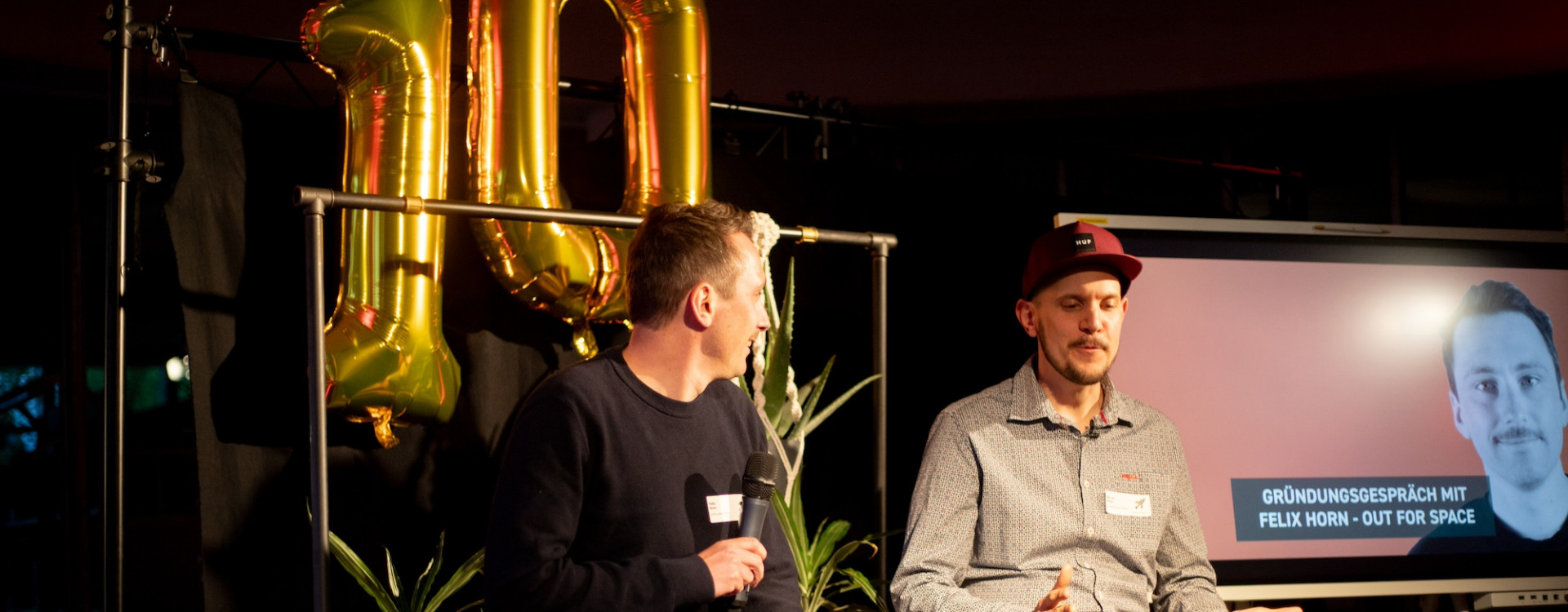 Zwei Männer sitzen auf einer Bühne vor einem schwarzen Vorhang