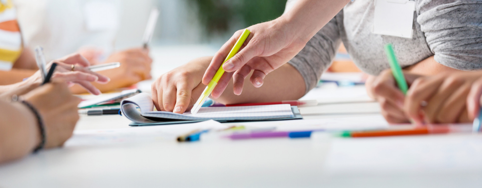 Schreibtisch mit Zetteln, bunten Siften und arbeitenden Personen