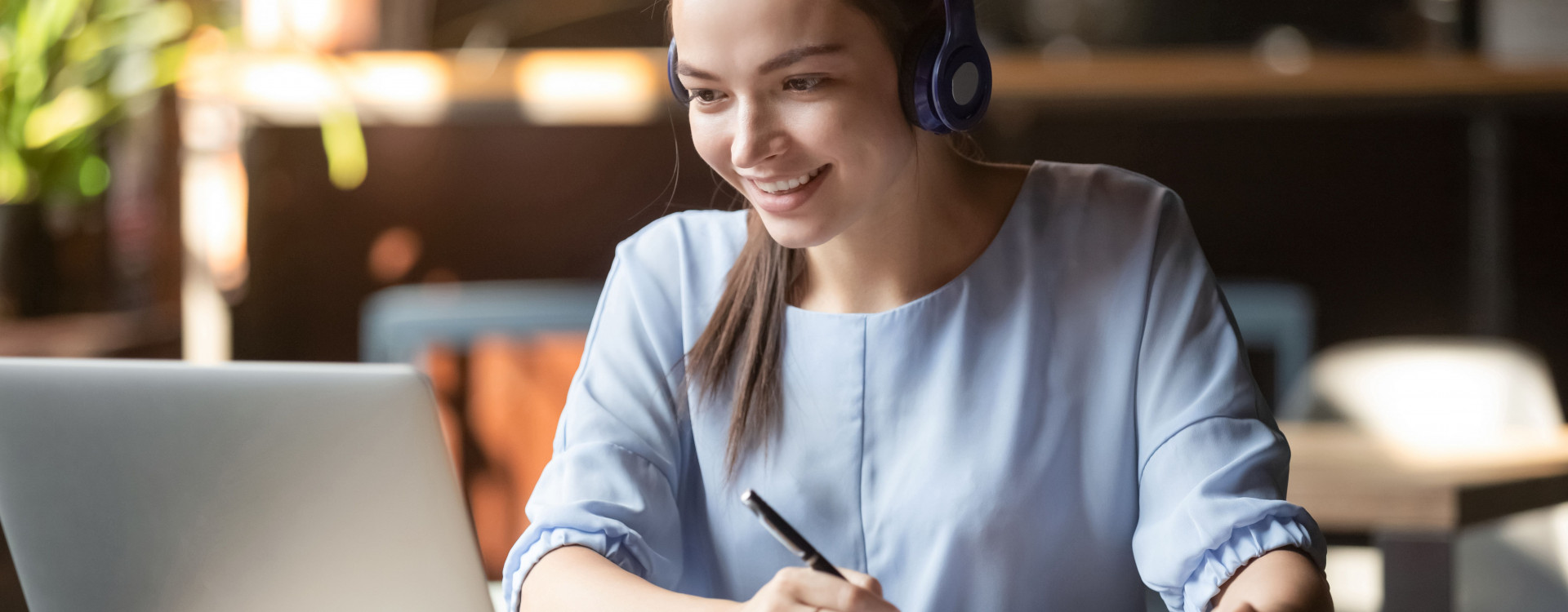Auf dem Bild ist sitzt eine weibliche Studierende mit Headset vor einem Laptop und macht Notizen auf einem Papier wärend sie an einem Online-Seminar teilnimmt.