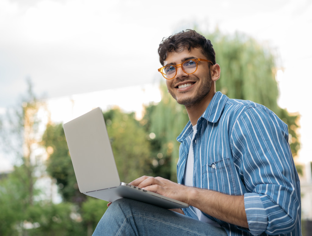 Ein Student sitzt in der Natur vor seinem Laptop. Er trägt eine Brille und ein blau gestreiftes Hemd über einem weißen T‑Shirt.