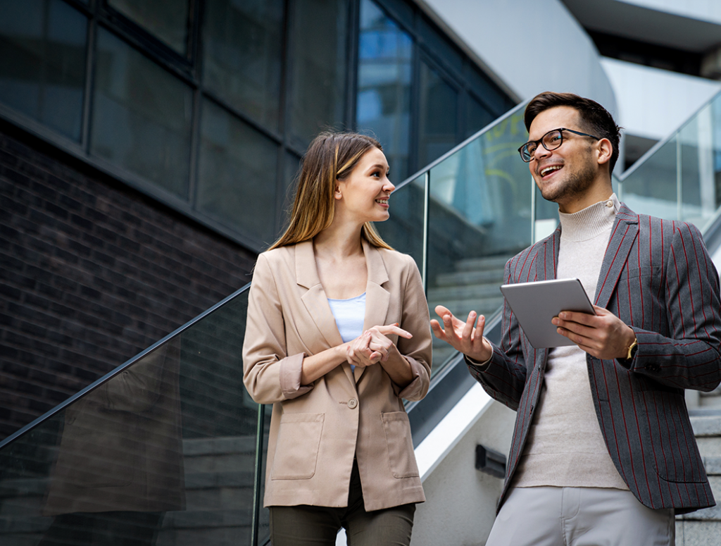 Eine Frau und ein Mann mit Tablet in der Hand, beide im Business Look, stehen auf einer Treppe in einem modernen Unternehmen und unterhalten sich,