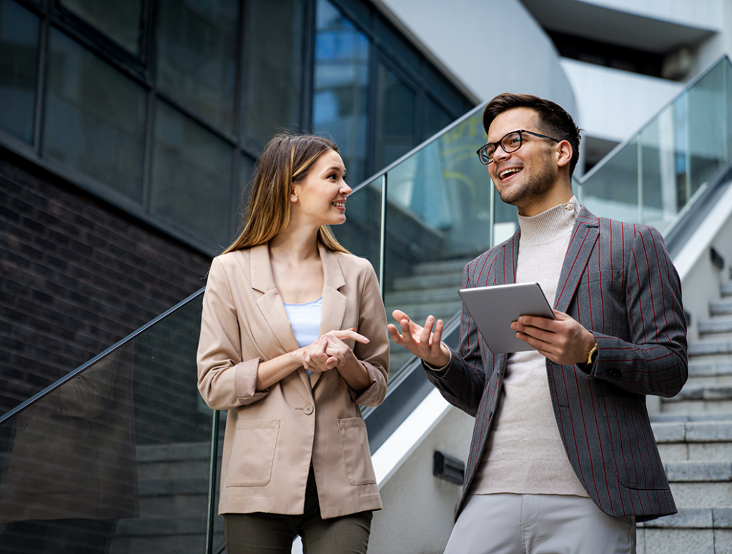 Eine Frau und ein Mann mit Tablet, beide im Business Look, stehen auf einer Treppe in einem modernen Unternehmen und unterhalten sich