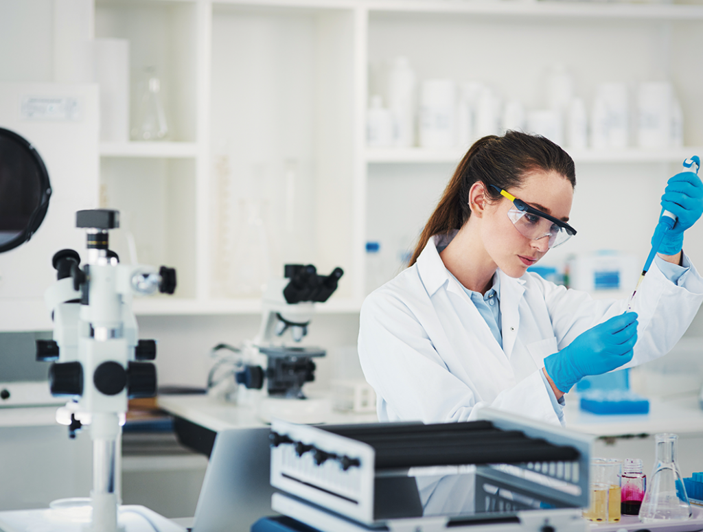 Eine Frau im weißen Kittel mit Schutzbrille und blauen Handschuhen steht in einem modernen Labor und arbeitet mit einer Pipette