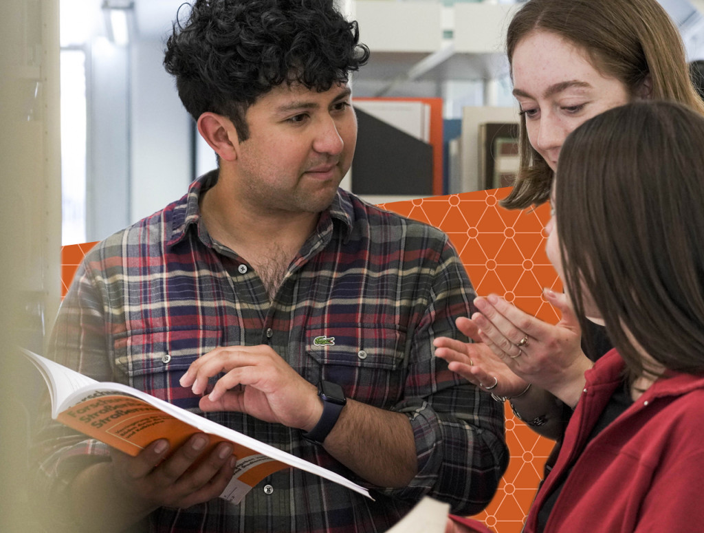 Drei Studierende stehen in Bibliothek und schauen in zwei Bücher