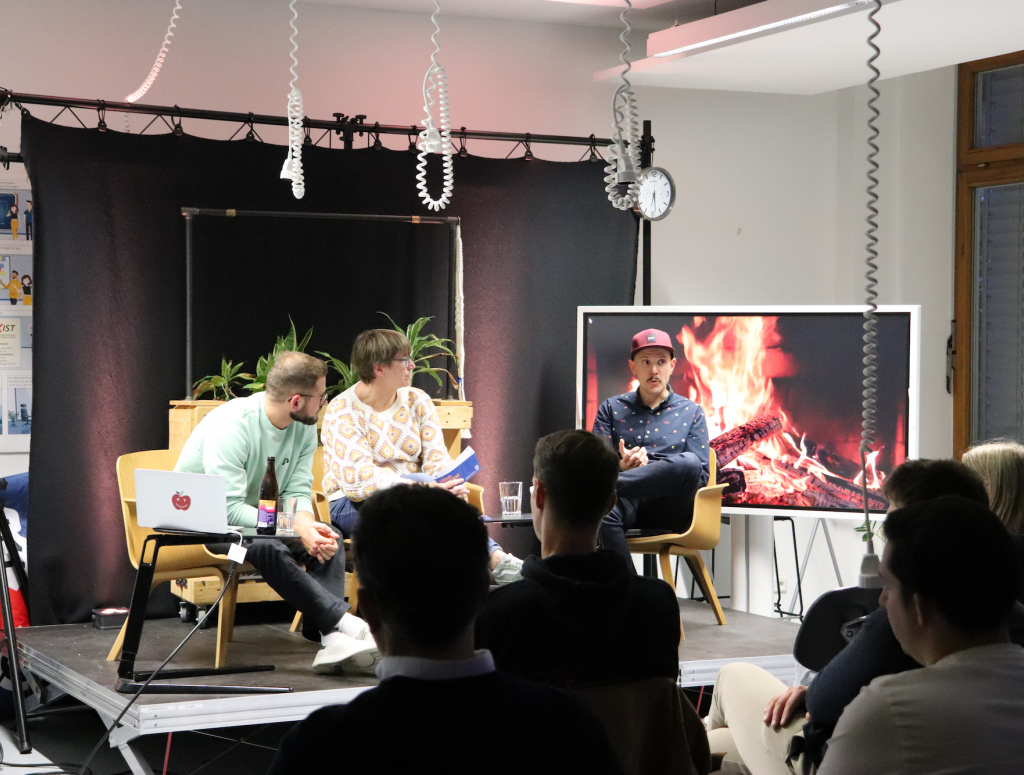 Personen sitzen auf einer Bühne. Im Hintergrund steht ein Bildschirm mit Kaminfeuer.