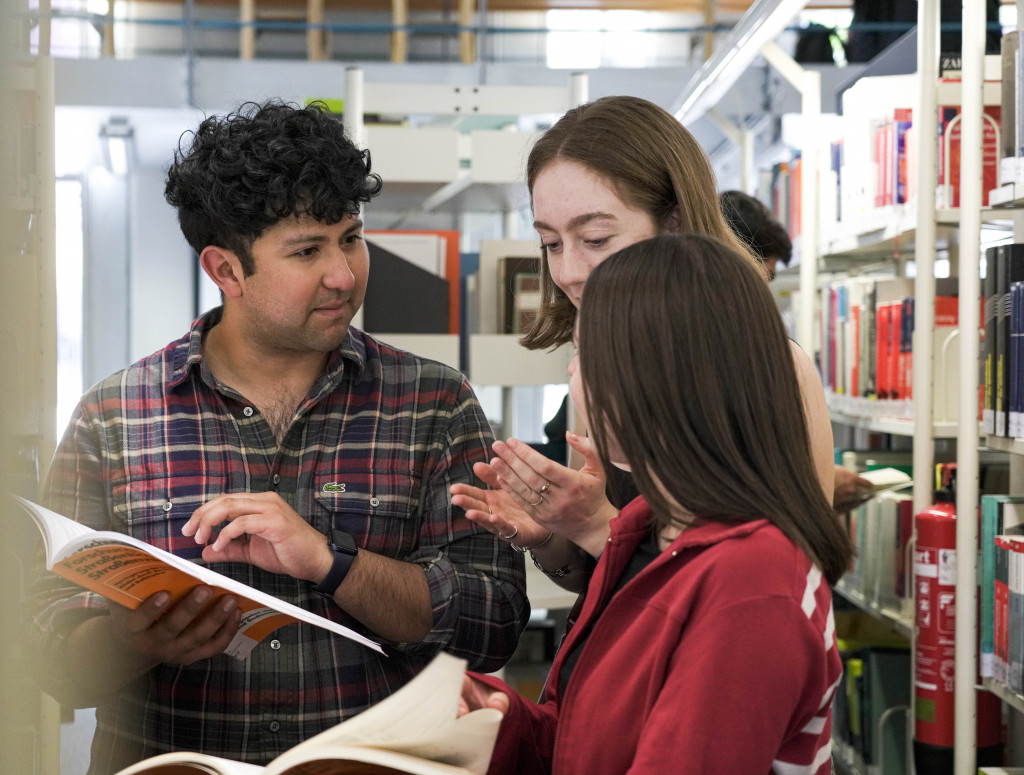 Drei Studierende stehen in Bibliothek und schauen in zwei Bücher