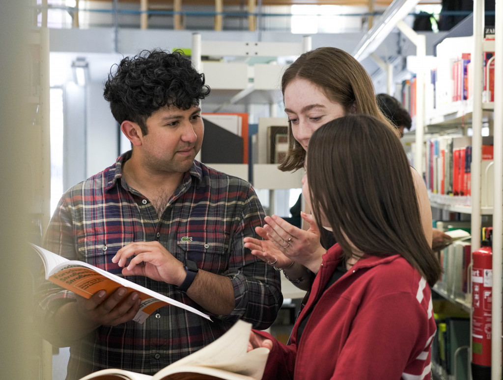 Drei Studierende stehen in Bibliothek und schauen in zwei Bücher
