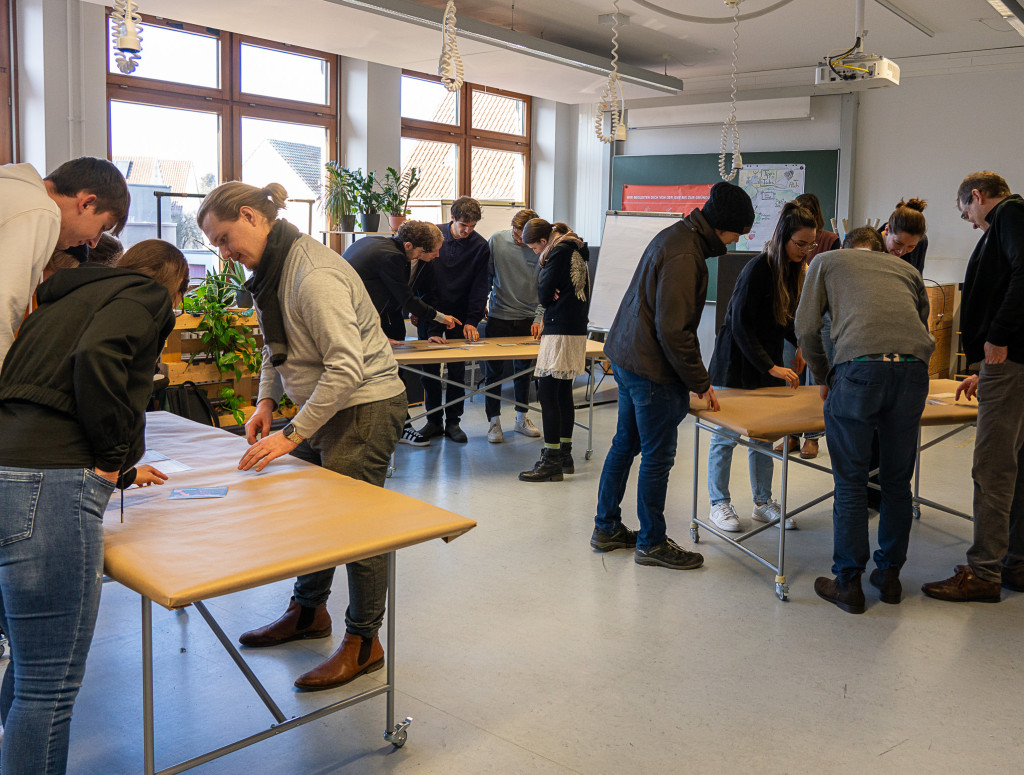 Personen stehen um drei Tische herum und arbeiten interaktiv