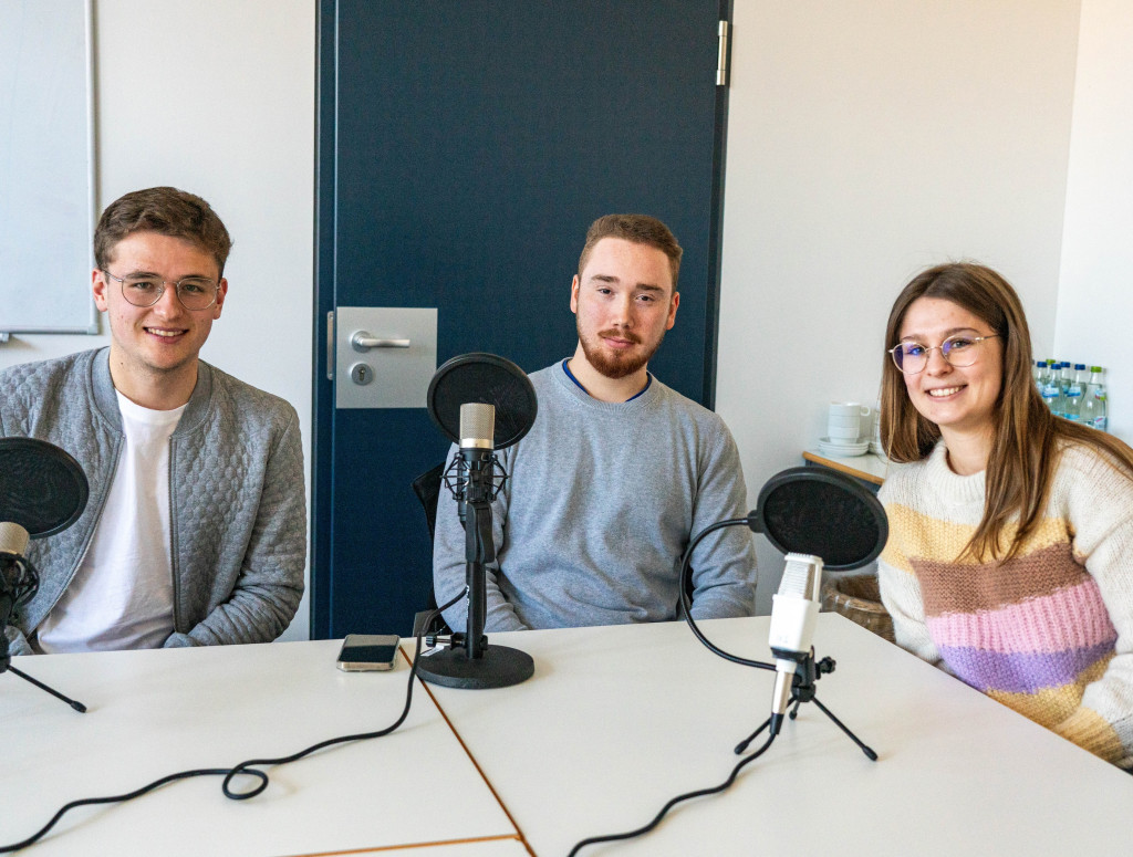 Drei Studierende sitzen mit Mikrofonen um einen Tisch