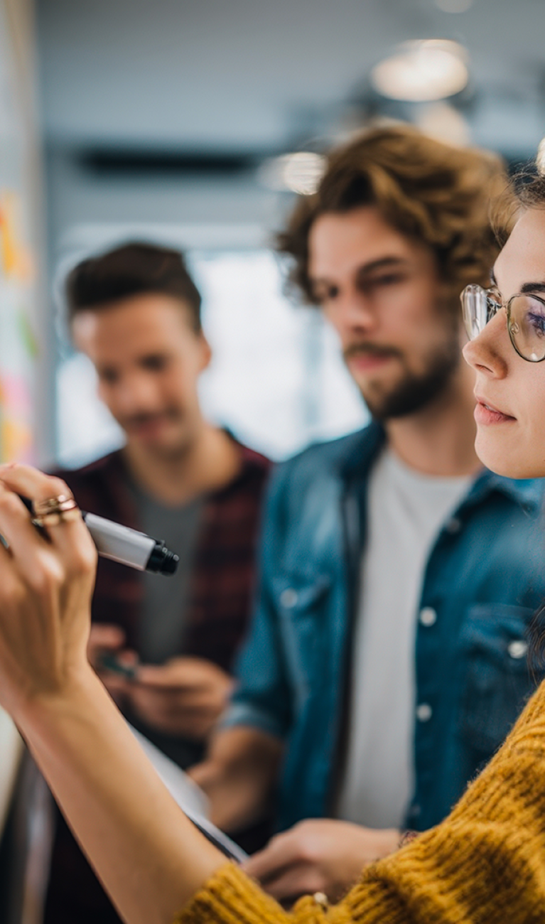 Studierende im legerem casual Look stehen mit Stift vor einem Whiteboard mit vielen bunten Post-its
