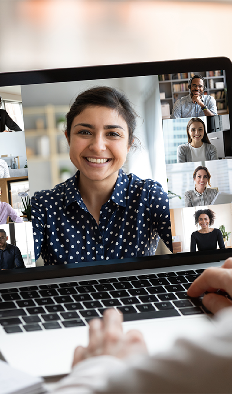 Frau mit weißer Bluse sitzt vor einem Laptop am Schreibtisch und nimmt an einem Online-Meeting teil, dessen Teilnehmer auf dem Bildschirm zu sehen sind