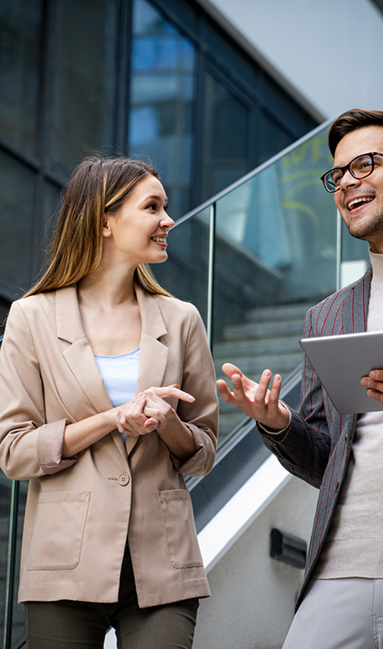 Eine Frau und ein Mann mit Tablet, beide im Business Look, stehen auf einer Treppe in einem modernen Unternehmen und unterhalten sich