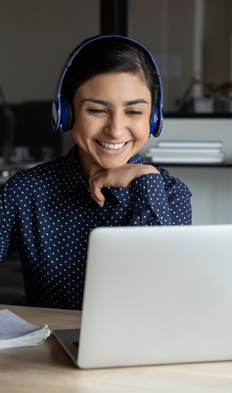 Junge Frau sitzt lächelnd it Headset und Schreibset vor einem Laptop in einem modernen Büro