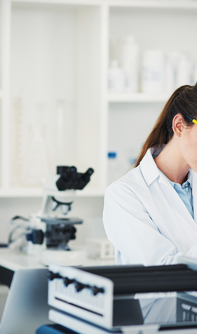 Eine Frau im weißen Kittel mit Schutzbrille und blauen Handschuhen steht in einem modernen Labor und arbeitet mit einer Pipette