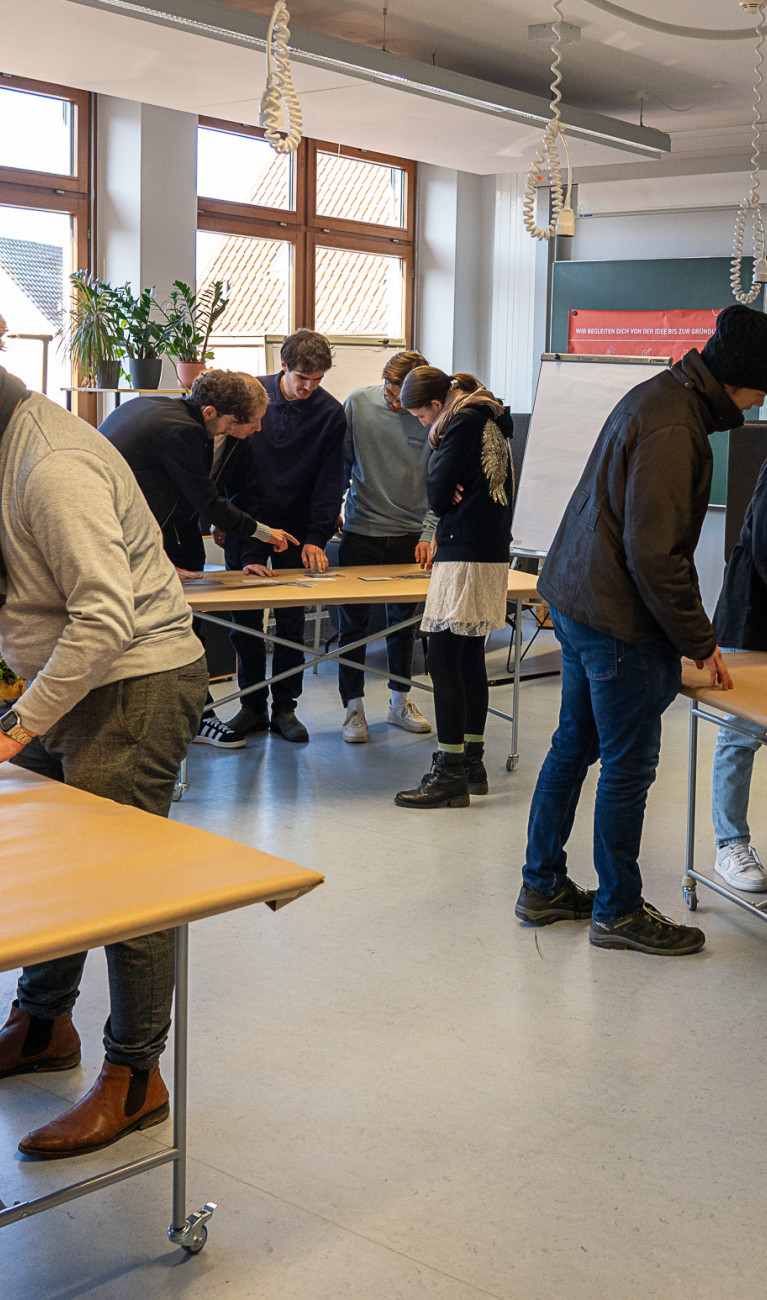 Personen stehen um drei Tische herum und arbeiten interaktiv