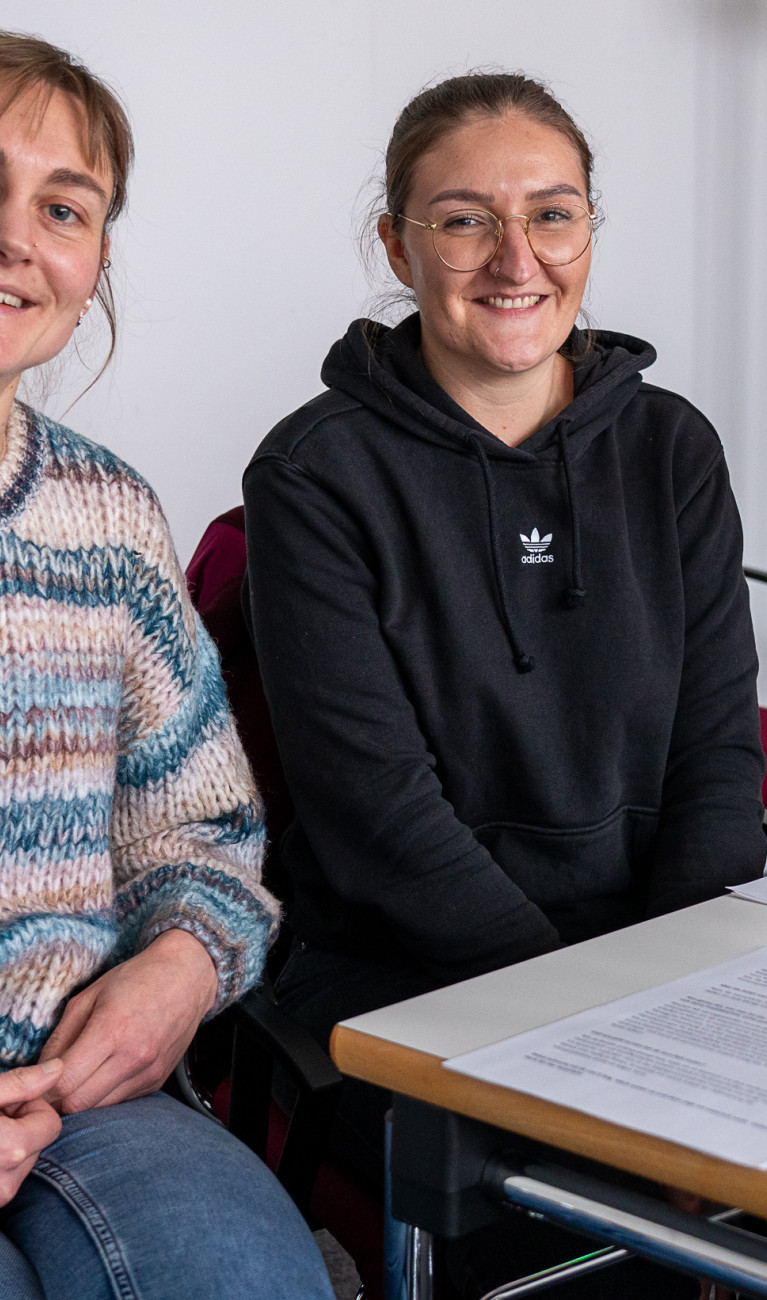 Zwei Frauen sitzen an einem Tisch bei einer Podcastaufnahme und lächeln in die Kamera