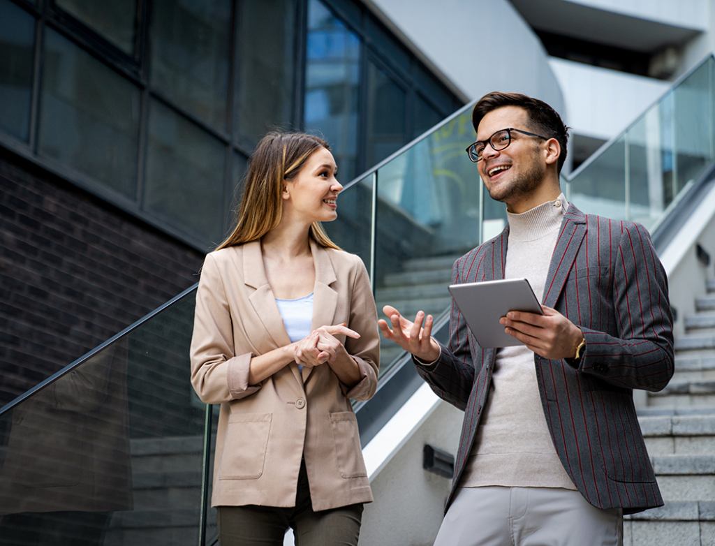 Eine Frau und ein Mann mit Tablet, beide im Business Look, stehen auf einer Treppe in einem modernen Unternehmen und unterhalten sich