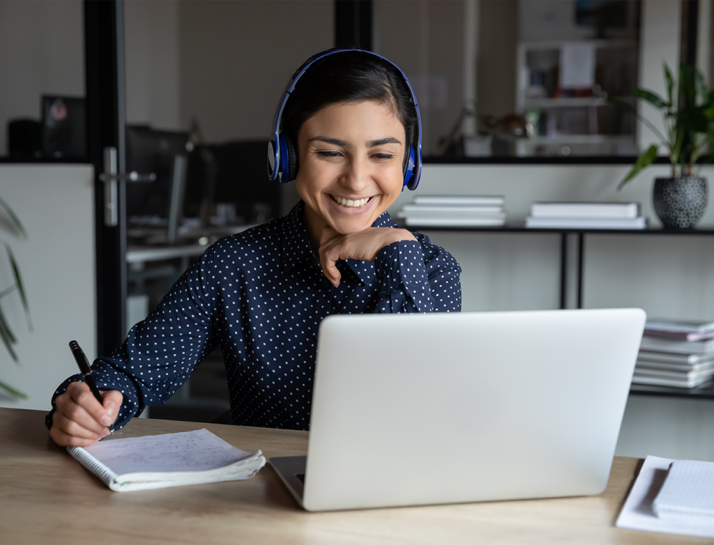 Junge Frau sitzt lächelnd it Headset und Schreibset vor einem Laptop in einem modernen Büro