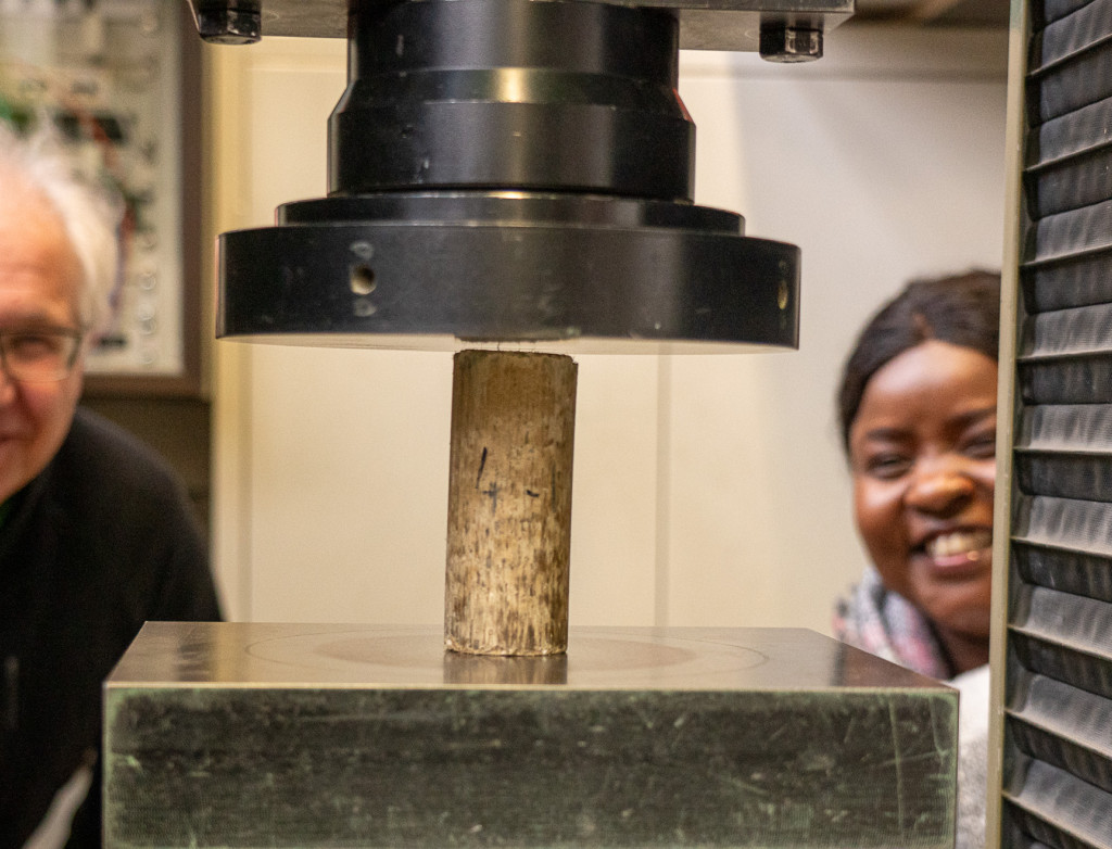 Zwei Personen schauen sich einen Lehmstein ein, der von einer Maschine zusammengepresst wird