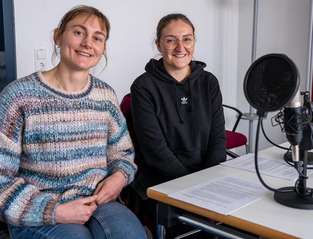 Zwei Frauen sitzen an einem Tisch bei einer Podcastaufnahme und lächeln in die Kamera