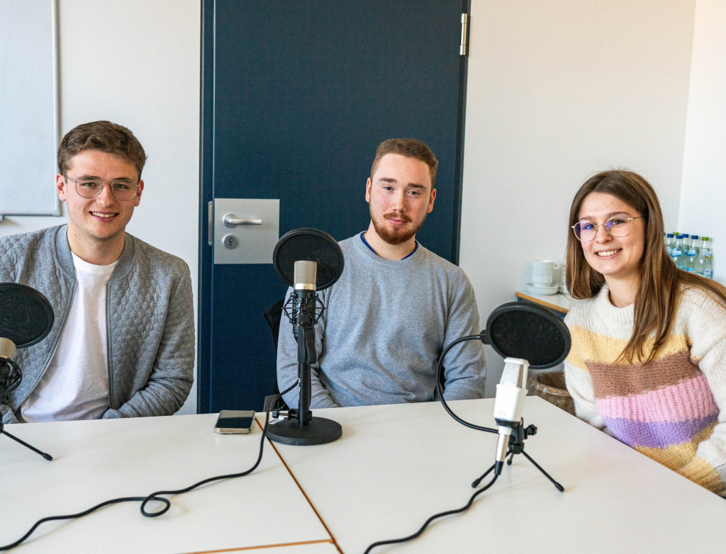 Drei Studierende sitzen mit Mikrofonen um einen Tisch