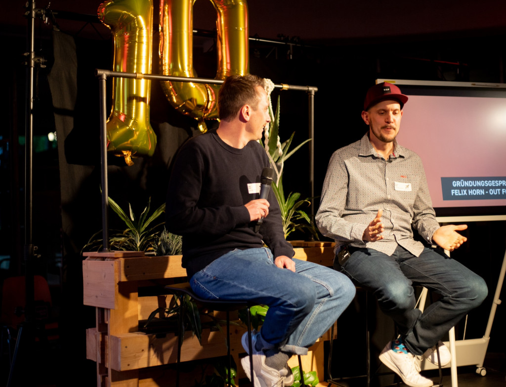 Zwei Männer sitzen auf einer Bühne vor einem schwarzen Vorhang